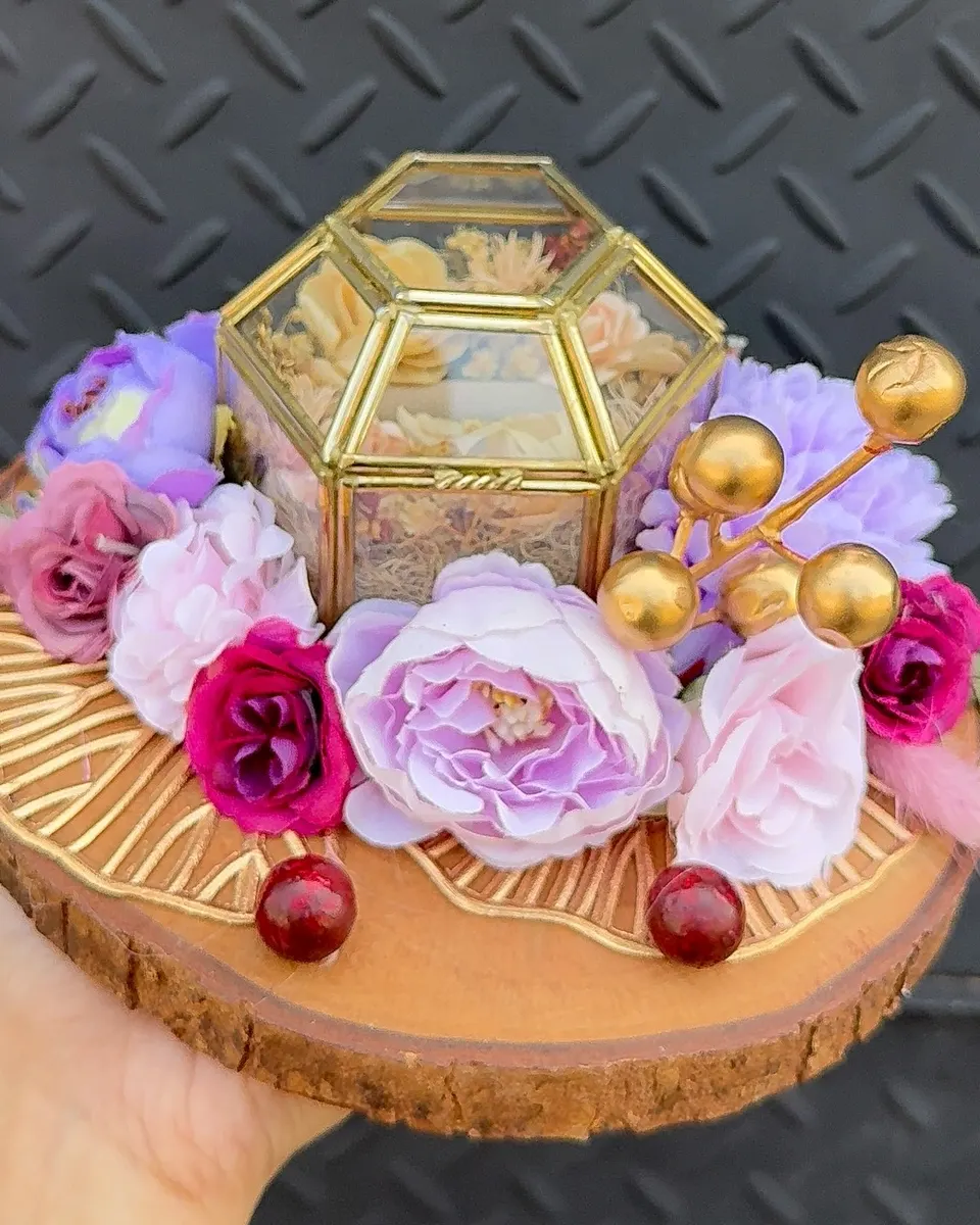 Ring box kaca emas di atas potongan kayu, dikelilingi mawar ungu lilac dan pink