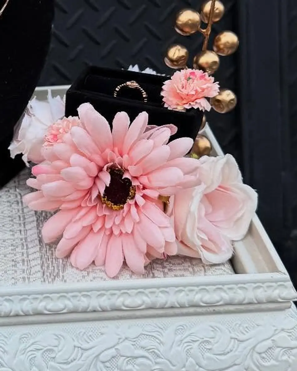 Kotak cincin beludru hitam berisi cincin, dengan gerbera pink dan buah hias emas