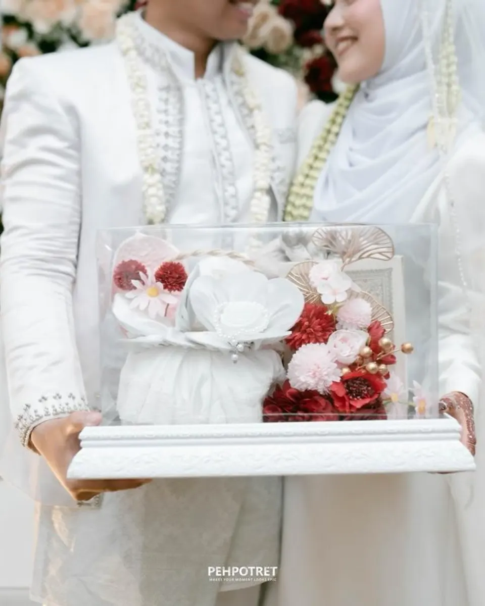 Pengantin membawa kotak akrilik bening berisi kain putih dan bunga merah di atas nampan ukir putih
