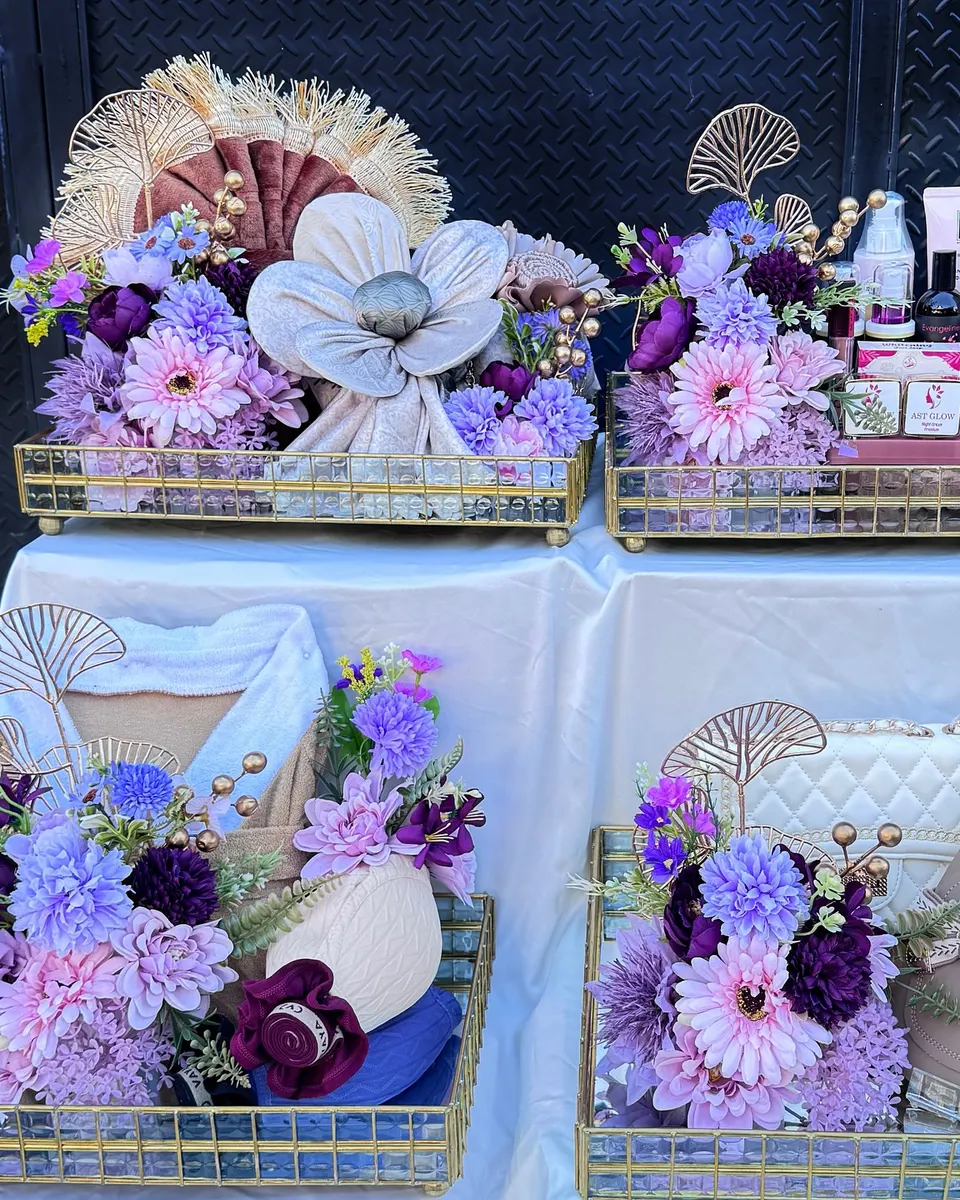 Empat nampan cermin emas berisi kain dibentuk bunga, kosmetik, tas, dan sandal, dengan bunga ungu lilac